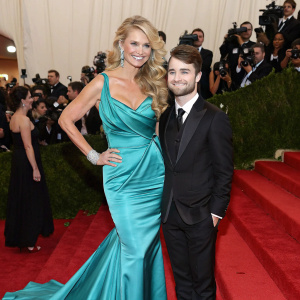 Christie Brinkley and Daniel Radcliffe at the Met Gala. Christie in a stunning turquoise silk gown, Daniel in a tailored black suit. Crowded red carpet scene with flashing cameras.