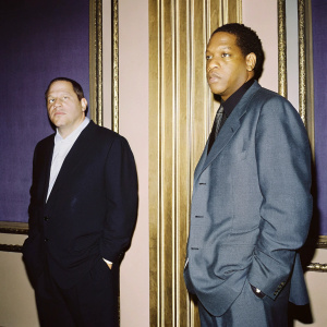 Harvey Weinstein and Jay-Z in a formal gathering. Dressed in dark suits. Serious expressions. Purple backdrop with gold accents.