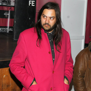 Photograph of Kurt Cobain. Long dark hair, facial hair, intense gaze. Festival stage background