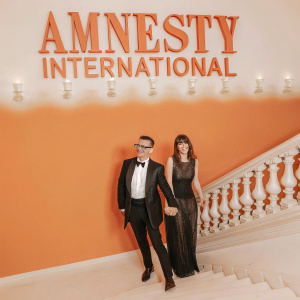 Fred Armisen and Eleanor Friedberger at a high-profile gala. Wearing formal attire. They are standing beside a large banner that reads 'Amnesty International'.
