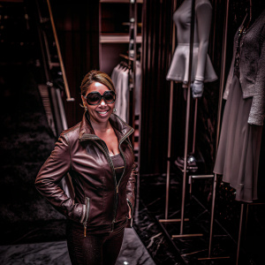 Mariah Carey, wearing a brown leather jacket and sunglasses, posing in a boutique. White garment on a mannequin in the background.