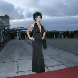 Kym Marsh on a bustling red carpet. Stylish black gown with deep V-neckline. Bob haircut with voluminous texture. Warm smile. Crowd and photographers in the background