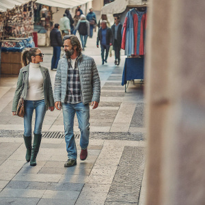 Andrea Pirlo and Valentina Baldini strolling through a bustling city market. Andrea, bearded and in sunglasses, dons a plaid shirt and jeans. Valentina, with a ponytail and boots, wears a quilted jacket over a white top. Vibrant street scene with colorful stalls and shoppers.