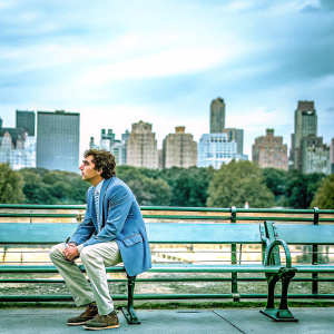 Larry David, dressed in a blue blazer and khakis, sitting on a park bench. A city skyline backdrop.