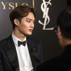 Kai from EXO in a formal interview setting. Dark backdrop with YSL logo. Elegant attire.