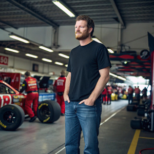 Dale Earnhardt Jr., dressed in a casual black t-shirt and jeans, standing in a NASCAR garage. The scene is vibrant with racing equipment and crew members in the background.
