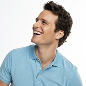 Jonathan Groff, actor, smiling warmly. Wearing light blue polo shirt. White studio background