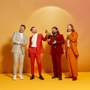 Marcus Mumford, Ben Lovett, Winston Marshall, and Ted Dwane, dressed in tuxedos, at the Grammy Awards. Crowd cheering