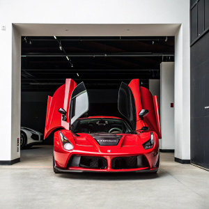 A red Ferrari LaFerrari supercar with black trim, parked in a garage with white walls. Open doors reveal intricate engine. Black background