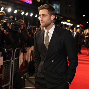 Oliver Jackson-Cohen on the set of a film premiere, dressed in a classic black suit with a subtle, textured tie. Crowded event, flashing cameras.