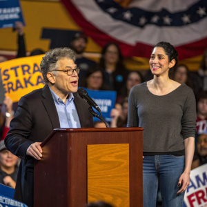 Political rally featuring Senator Al Franken and comedian Sarah Silverman. Sen Franken delivers speech at podium, Sarah smiles from the side.