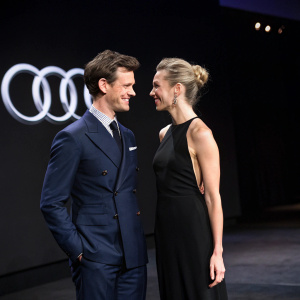 Matt Smith and Vanessa Kirby attend the unveiling of the new Audi model. Matt Smith in a navy suit, friendly smile. Vanessa Kirby in a sleek black gown, bun hairstyle. On a dimly lit stage with an Audi logo backdrop.