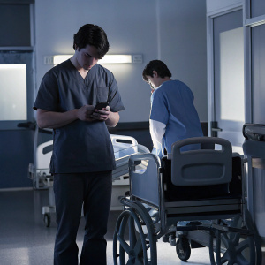 Finn Wittrock and an extra from 'All My Children' in a hospital scene. Finn checks his phone while the extra stands nearby. Both are in scrubs.