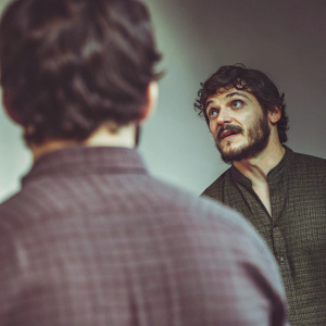 Pedro Pascal in a candid photo shoot. He sports a full beard and mustache, dark curly hair. Wearing a black textured shirt against a gradient backdrop.