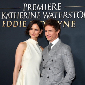 Katherine Waterston and Eddie Redmayne attending a glamorous premiere event. Katherine dons an elegant white, sleeveless gown, paired with a chic bob hairstyle. Eddie is impeccably dressed in a gray tailored suit with styled hair. Dark backdrop featuring the movie title.
