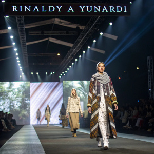 Rinaldy A Yunardi's fashion show. Models strut on the runway, showcasing unique, layered outfits. Bright lights and the designer's name displayed prominently.
