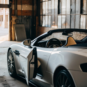 Aston Martin Vanquish Volante Carbon Edition parked in an open urban garage. Sleek white body with black accents. Visible interior is black. Convertible top down. Large garage door opens onto city street.