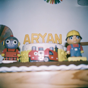 Birthday cake featuring characters from Chuggington and Bob the Builder. 'Aryan' written in playful font. Simple white backdrop