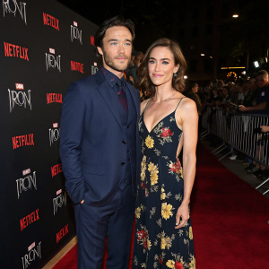 Tom Pelphrey and Jessica Stroup attend a Marvel event for 'Iron Fist'. Tom in a navy suit, Jessica in a floral maxi dress. Dark background with red carpet and fans
