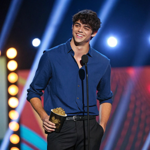 Noah Centineo, youthful actor, at award show. Dark hair, wearing blue shirt, black pants. Smiling broadly. Bright stage lights and vibrant backdrop behind him.