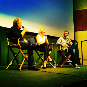 Sir Ridley Scott, Charlize Theron, and Michael Fassbender seated on stage at a Q&A session. Bright studio lights and smiling audience visible