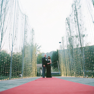 John Aniston and Sherry Rooney Aniston at the annual Hollywood Awards. John in a classic dark suit. Sherry in an elegant black gown. A vibrant red carpet backdrop with twinkling lights