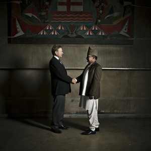 Formal handshake between Per Stig Møller, Danish Foreign Minister, and Pushpa Kamal Dahal, Nepalese Prime Minister. Against a vibrant mural backdrop depicting cultural symbols. Both in traditional attire.