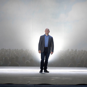Jeff Bezos, smiling broadly on stage. Formal attire, blue button-down shirt with a plaid blazer. Crowded auditorium, dimly lit background.