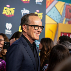 Clark Gregg, in a dark suit with glasses, at the Comic-Con event. Backdrop features logos and vibrant colors, attendees crowd around
