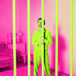 Black and white photograph of Roddy McDowall from The Twilight Zone. He is standing behind bars in a dimly lit room. The background features antique furniture and household items. The contrast and textures are emphasized.