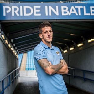 Stevan Jovetic, a professional footballer, in a casual blue polo shirt with tattoos visible, poses with a stern look in the tunnel at Etihad Stadium. The background showcases the stadium's seating and infrastructure along with a large 'Pride in Battle' banner.