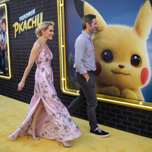Megyn Kelly and Douglas Brunt attend the grand premiere of Pokemon Detective Pikachu. Megyn in a floral-patterned pink gown, Douglas in casual attire. Walking in front of a massive, animated backdrop showcasing Pikachu.