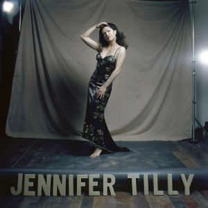 Jennifer Tilly, asian actress, in black evening gown with gold embroidery, posing gracefully. Soft studio lighting, plain beige backdrop, large font text below her name