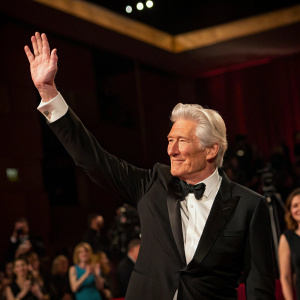 Richard Gere at a gala event, waving charmingly. Formal attire, dimly lit room, blurred audience in background.