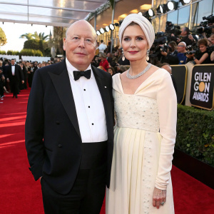 Julian Fellowes and Emma Fellowes on the Golden Globe Awards red carpet. Julian in a classic tuxedo. Emma in an elegant white gown with a matching headscarf. Crowd and photographers in the background.