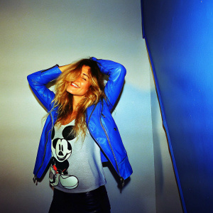 Alejandra Onieva, with her big blonde waves and a joyful smile, poses in a Mickey Mouse shirt and blue leather jacket. Shot in an empty studio with white backdrop and Madrid logo.