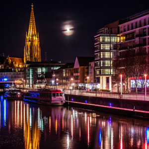 A stunning night view of Regensburg, Germany. The towering spire of the Regensburg Cathedral stands out under the moonlit sky, casting a golden glow. The river reflects the city's lights and buildings, creating a mirror-like image. The cityscape blends modern and historical architecture, illuminated by a mix of warm and cool tones, capturing a realistic and enchanting scene.