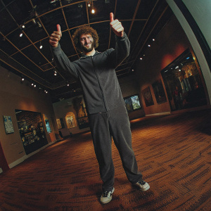 Lil Dicky inside the Grammy Museum, giving a thumbs up. Casual attire, bright indoor setting.