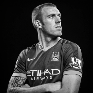 Richard Dunne, soccer player, serious face, short hair, tattoo visible. Wearing a sponsored jersey with airline logo on a plain black background.