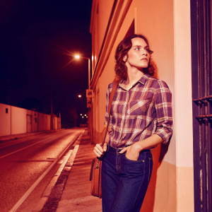 Daisy Ridley, in a casual plaid shirt and jeans, stands alone on a deserted street at night. She looks contemplative and serene. The urban backdrop is quiet and peaceful.