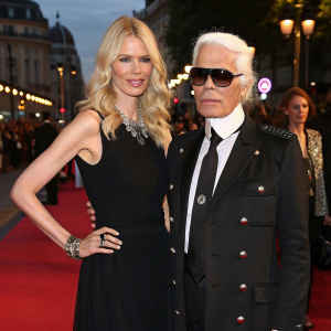 Karl Lagerfeld and Claudia Schiffer at a high-profile fashion gala. Claudia wears a black sleeveless dress, Karl in a black military-style jacket and black tie. The backdrop is a bustling street scene.
