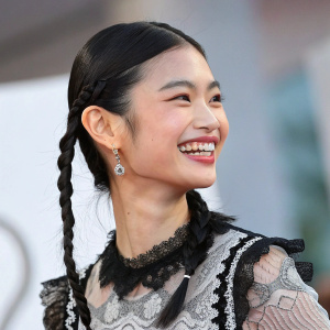 Close-up of HoYeon Jung, Asian actress, with radiant smile. Dark straight hair with braids, youthful look. Wearing black and gray outfit with lace details. Blurred background