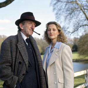 Ron Moody and Cassie Yates from 'Nobody's Perfect' posing outdoors. Moody in a dark hat and tweed coat, smoking a pipe. Yates in a light blazer and blouse.