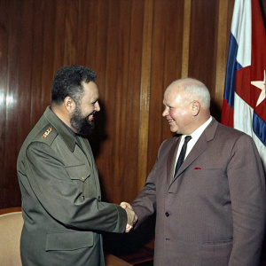 Fidel Castro and Nikita Khrushchev shaking hands during a diplomatic meeting. They are in a formal, wood-paneled conference room with a large flag hanging on the wall behind them.