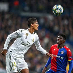 Raphael Varane of Real Madrid and Breel Embolo of FC Basel in intense match play. Varane in white jersey with Fly Emirates logo. Embolo in blue jersey with European Champions League emblem. Ball in mid-air between them. Outdoor stadium packed with spectators.