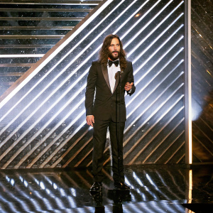 Jared Leto accepting a prize at the Screen Actors Guild Awards. Dressed in a formal black tuxedo, long wavy brown hair. Shimmering backdrop with dramatic lighting.