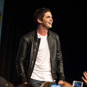 Logan Lerman at Comic-Con. Wearing a black leather jacket and a white t-shirt. Dark background, surrounded by eager fans.