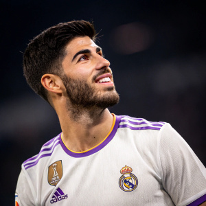 Close-up portrait of Marco Asensio. Groomed beard, white and purple soccer jersey with team logos. Dark, blurred background.