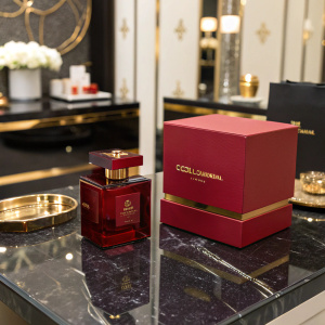 Dolce & Gabbana's 'The One' fragrance displayed on a sleek black marble countertop. Deep red bottle with gold accents, sitting in a matching box, luxury event setup