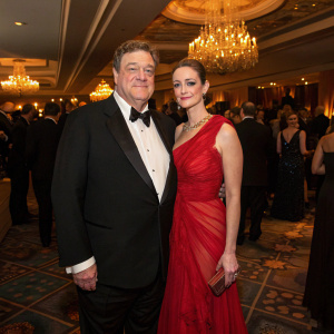 John Goodman and Annabeth Hartzog attending a gala. John in a black tuxedo, Annabeth in a flowing red gown. Crowded room with chandeliers.